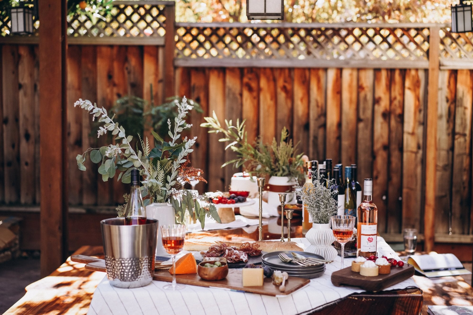 A long outdoor table set with wine, cheese and greenery
